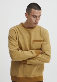 Sudadera de color mostaza hecha de un tejido suave, con un cuello redondo acanalado y un pequeño bolsillo delantero con un borde en contraste.
