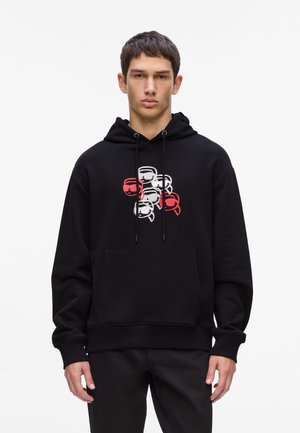 Schwarzer Kapuzenpullover mit Kängurutasche, ausgestattet mit einem mehrfarbigen Grafikdesign von Gesichtern in Rot, Weiß und Schwarz auf der Brust. Mit Kordelzug an der Kapuze.
