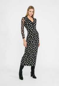 Sort midi-kjole med polka dots, der har gennemsigtige, mønstrede lange ærmer og krave, parret med knæhøje sorte støvler.