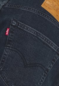 Jeans de denim azul oscuro, con una etiqueta de cuero, un tab rojo y una costura curva distintiva en el bolsillo trasero. Vista en primer plano.