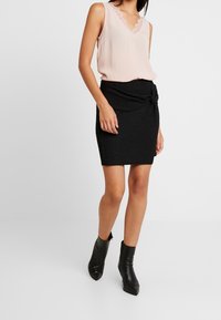 Kvinna som bär en ärmlös ljusrosa topp med spetsdetaljer, en svart texturerad omlottkjol och svarta spetsiga ankelskålar, stående framför vit bakgrund.