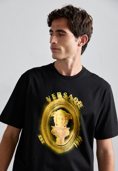 Young man wearing a black T-shirt with a gold Medusa emblem and "Versace Est 1978" text, looking to his left against a plain background.