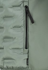 Grüne gesteppte Jacke mit schwarzem vertikalem Reißverschluss und markiertem Zugband auf der linken Seite, mit einem kleinen schwarzen Stoffetikett am unteren Ende.