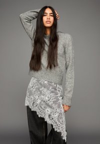 Žena s dlhými tmavými vlasmi, oblečená v šedej pletenej svetri, asymetrickej bielej čipkovej sukni cez tmavé široké nohavice, pózuje na sivom pozadí.