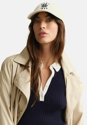 Vrouw met lang bruin haar, die een beige pet met embleem draagt, een beige trenchcoat en een marineblauw geribd shirt met witte kraag en knopen.