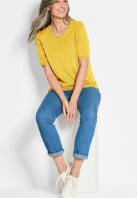 Gelbes kurzärmliges Strickoberteil mit rundem Halsausschnitt, kombiniert mit aufgerollten blauen Denim-Jeans und weißen Sneakers. Sie sitzt auf einem weißen Sockel.