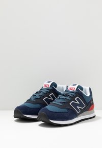 Pár tmavě modrých a šedých tenisek New Balance 574 s bílými a rudými akcenty, umístěný vedle sebe na bílé ploše.