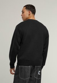 Svart stickad tröja sedd bakifrån, med ribbade mudd och nederkant. I kombination med mörka jeans som visar kontrasterande sömdetaljer.