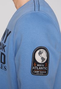 Felpa blu in morbido tessuto con testo stampatonero e una toppa rotonda con un design di globo e dettagli in arancione e bianco.