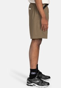 Braune Shorts mit elastischem Bund und Seitentasche, kombiniert mit schwarzen Sportschuhen und schwarzen Socken, dargestellt in einer Frontalansicht von der Seite.