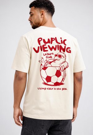 Mann trägt beige T-Shirt mit roter Grafik eines entspannten Vogels in einem Fußball und Text "Public Viewing Leisure Club Living easy is the goal"