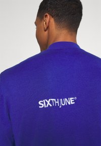Lila stickad tröja med hög krage, med vit broderad logotyp "SIXTHJUNE" på baksidan. Slät textur, åtsittande design.