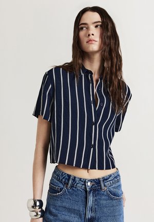 Jeune femme aux cheveux bruns longs portant un haut court à manches courtes rayé verticalement bleu marine et blanc, ainsi qu'un jean bleu taille haute.