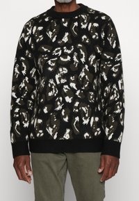 Pull à motifs noirs, verts et blancs avec un col rond, des manches longues et des poignets et ourlet côtelés. Tissu texturé avec un design abstrait.