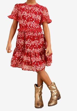 Barn iført en rød kjole med lyserøde hjerte mønstre og flæse detaljer, kombineret med skinnende gyldne cowboystøvler, står med krydsede ben.