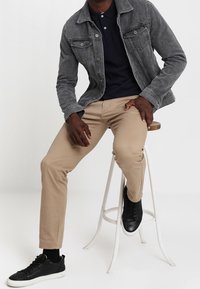 Grijze denimjack, marineblauw polohemd, tan chino's en zwarte sneakers. Zittend op een witte kruk, met de benen gekruist en de handen op de knie.