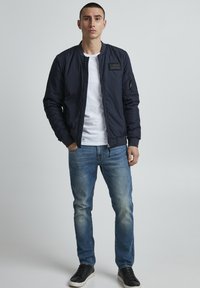 Chaqueta bomber azul marino con cremallera frontal, puños acanalados y parche horizontal; combinada con jeans de lavado claro y zapatillas negras.