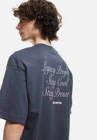 Camiseta oversized de color azul marino oscuro hecha de algodón, con gráficos en texto blanco en la parte posterior, que incluyen "Proyecto Legado" y "Mantente Fresco Mantente Presente."