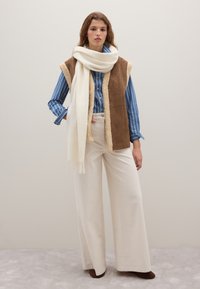 Gilet in finta pelliccia marrone con interno avorio, camicia a righe blu con bottoni, pantaloni bianchi a gamba larga e grande sciarpa bianca. Calzature marroni.