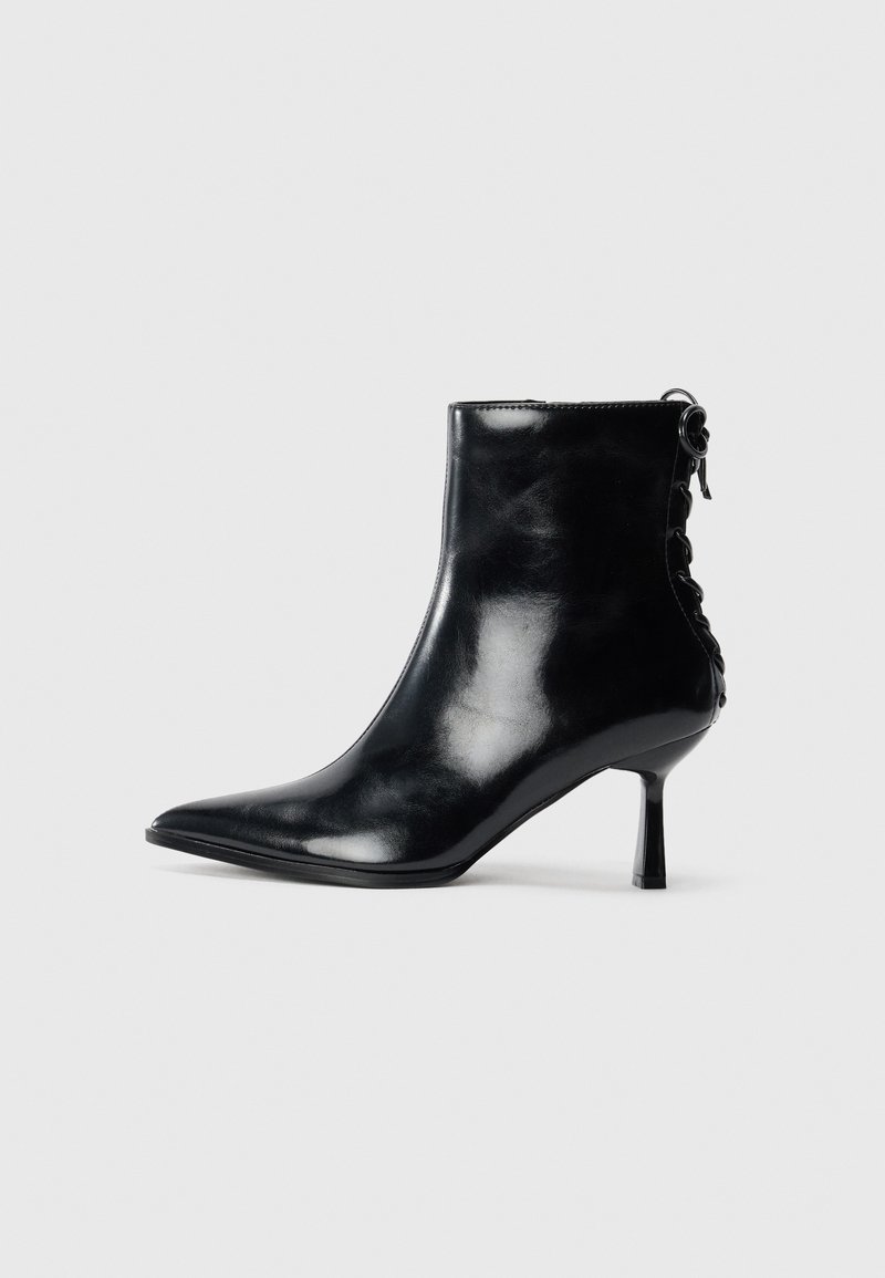Bottine en cuir noir brillant avec bout pointu, talon fin de hauteur moyenne et détail lacé à l'arrière sur un fond uni et clair.