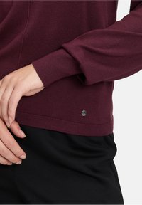 Burgunderfarbener gestrickter Pullover mit gerippten Bündchen und Saum. Mit einem kleinen metallischen Logozusatz in der Nähe des Saums. Glatte Textur, taillierte Passform.