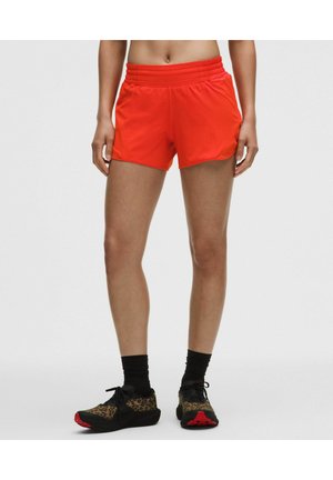Vrouw draagt feloranje sportshorts, zwarte sokken en zwarte hardloopschoenen met luipaardprint en rode zolen, staand tegen een effen achtergrond.