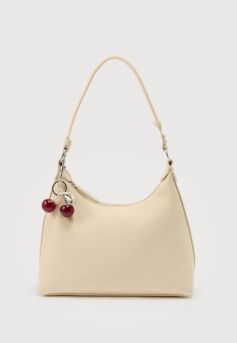 Sac à main de couleur crème avec surface texturée, une bandoulière unique, et un charm en forme de cerise sur tirette, sur un fond uni.