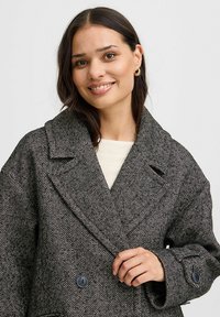 Šedý kabát s potlačou houndstooth, ktorý má dvojradové zapínanie, veľké klopy a dva gombíky, vyrobený z textúrovanej látky.
