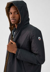 Chaqueta con capucha negra de material exterior suave y un interior marrón en contraste. Presenta puños elásticos y un parche de logo en la manga.