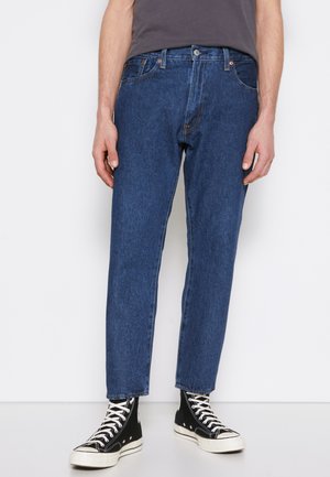 Jeans en denim bleu avec un design à jambe droite, comportant cinq poches, une fermeture à bouton et des coutures contrastées. Portés avec des baskets montantes noires.