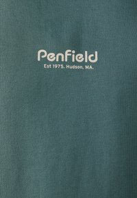 Žalias medvilnės audinys su briaunotu raštu, kurio centre yra spaudinys balta spalva: „PenField Est 1975. Hudson, MA.“