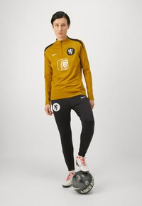 Nike treningsklær med en gull glidelåsjakke og svarte bukser, med logoer og en svart-hvitt fotball plassert på bakken.