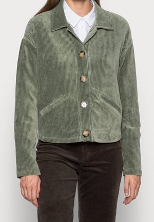 Persona que lleva una chaqueta de pana verde con grandes botones sobre una camisa blanca con cuello y pantalones marrón oscuro.