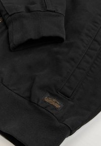 Gros plan sur un tissu noir montrant une manche de veste avec un poignet côtelé et une petite étiquette cousue avec un texte et un symbole de couronne.