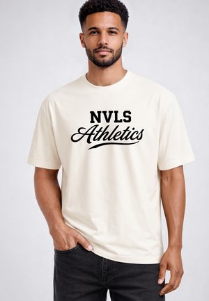 Man with short curly hair and beard wearing a white t-shirt with black text "NVLS Athletics," standing against a plain light background.
