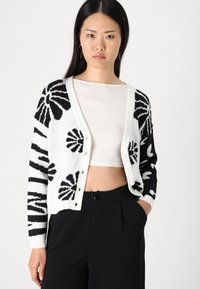 Black and white knitted cardigan featuring floral patterns, a v-neckline, button front closure, and ribbed cuffs, worn over a light top.