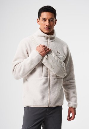 Homme portant une veste en polaire beige clair et un petit sac bandoulière beige, debout devant un fond uni.