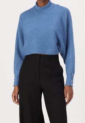 Kobieta ubrana w prążkowany, niebieski krótki sweter z mankietami na guziki oraz czarne, szerokie spodnie z wysokim stanem na jednolitym tle.