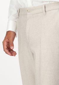 Beige maßgeschneiderte Hose aus strukturiertem Stoff, mit einem einzelnen Knopfverschluss, Gürtelschlaufen und einer Seitenhand im Bild.