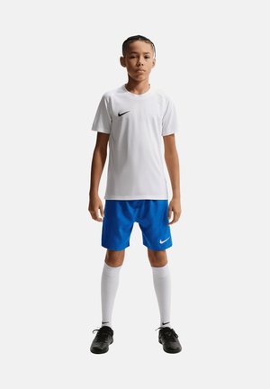 Ragazzo in piedi che indossa una maglietta sportiva bianca Nike, pantaloncini blu, calzini lunghi bianchi e scarpe da calcio nere su sfondo bianco neutro.