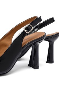 Décolleté in vernice nera con punta affilata, cinturino regolabile con fibbia e tacco medio a blocco. Sottopiede color marrone chiaro.