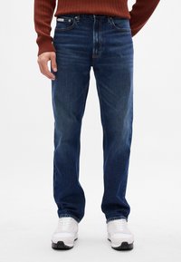 Jeans dritti blu scuro con lavaggio medio, design a cinque tasche e accessori in metallo, abbinati a scarpe da ginnastica bianche.