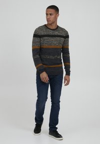 Maglione a righe in grigio scuro, crema e senape, con scollo a giro e polsini a costine, abbinato a jeans blu scuro e sneakers nere.