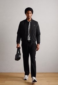 Jachetă bomber neagră cu manșete striate, tricou polo gri, pantaloni negri și adidași albi. Ține o geantă texturată neagră.