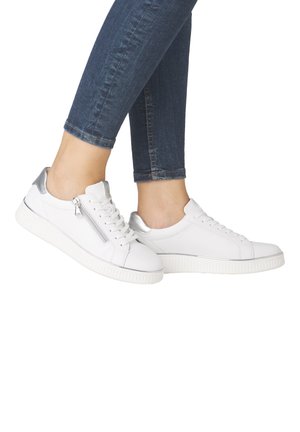 Personne portant des baskets basses blanches avec des accents argentés au talon et un détail de fermeture éclair, associées à un jean skinny bleu foncé.