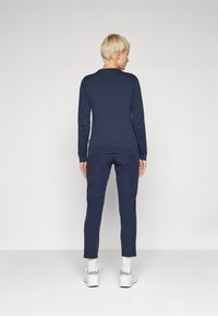 Sötétkék kötött pulóver hosszú ujjakkal, bordázott aljjal, hozzá illő sötétkék, slim-fit nadrággal és fehér sportcipővel viselve.