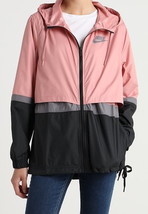 Kvinde iført en lyserød, grå og sort Nike hættejakke med forreste lynlås og blå jeans.