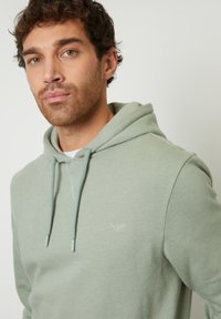 Grijs-groen hoodie met trekkoorden en een klein logo op de borst. Soepele textuur en een relaxte pasvorm, voorzien van een kangoeroezak.