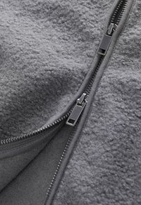 Tissu en polaire gris avec une surface texturée, doté d'une fermeture éclair robuste. L'intérieur est lisse et doublé, renforçant la chaleur et le confort.
