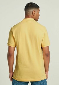 Polo giallo con una texture morbida, maniche corte e colletto. La vista dal retro mostra un orlo leggermente curvato e nessun motivo visibile.